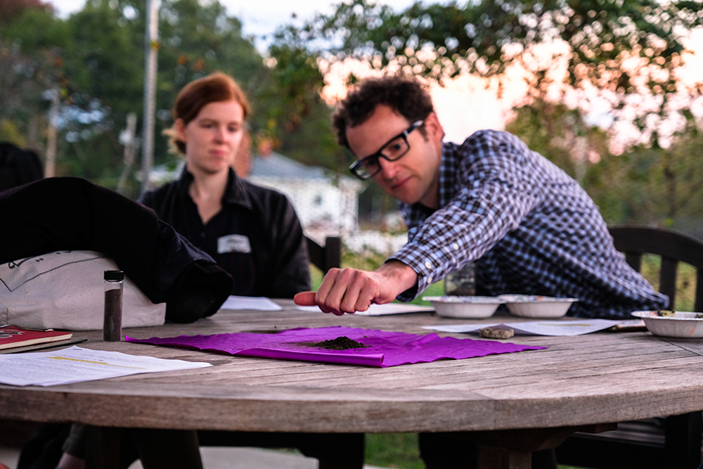 Two people seated outdoors at a table; one deposits a handful of dirt on to a cloth, while the other looks on.