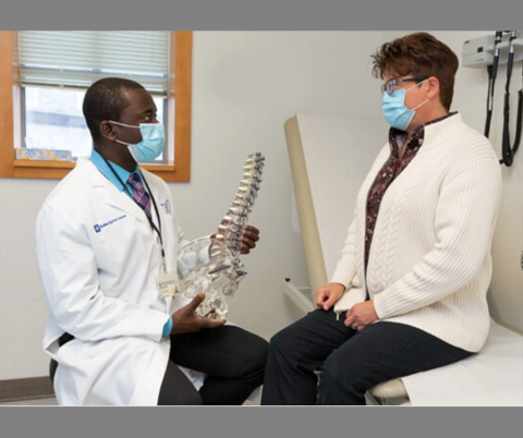 Male doctor holds model of spine and speaks with female patient seated on examining table.