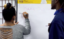 Woman viewed from behind writing on a large piece of easel paper under heading Baby Supplies, with man next to her.