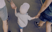 Small child seen from behind, walking while holding on to hands of adults on either side.