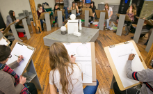 Students sit in a circle with easels, sketching an abstract sculpture as a teacher looks on.