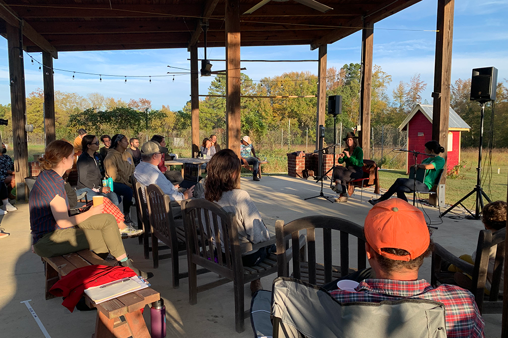 Outdoor scene of people seated and looking toward three presenters, who are also seated.