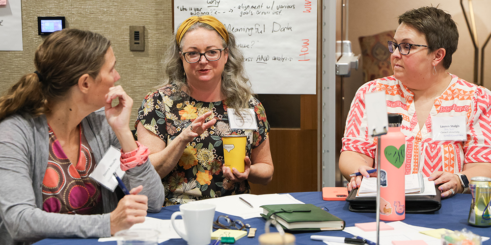 Three women engage in discussion while seated at a table.